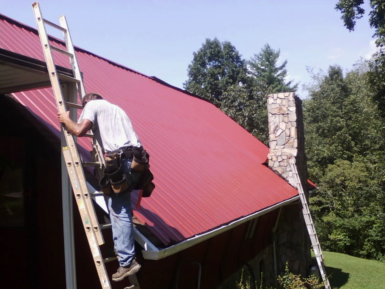 Roofer on a ladder beside a red metal roof and stone chimney — Soffit & Fascia Repair by Roofing Brothers in Abbottstown
